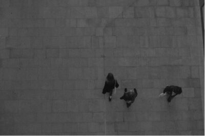 Dark grey photograph of three people wearing coats and holding bags, walking across a paved square. Photograph taken from above.